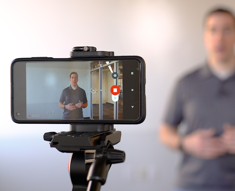 A videographer demonstrates how to use a cell phone and tripod equipment to take quality marketing videos for a school website.